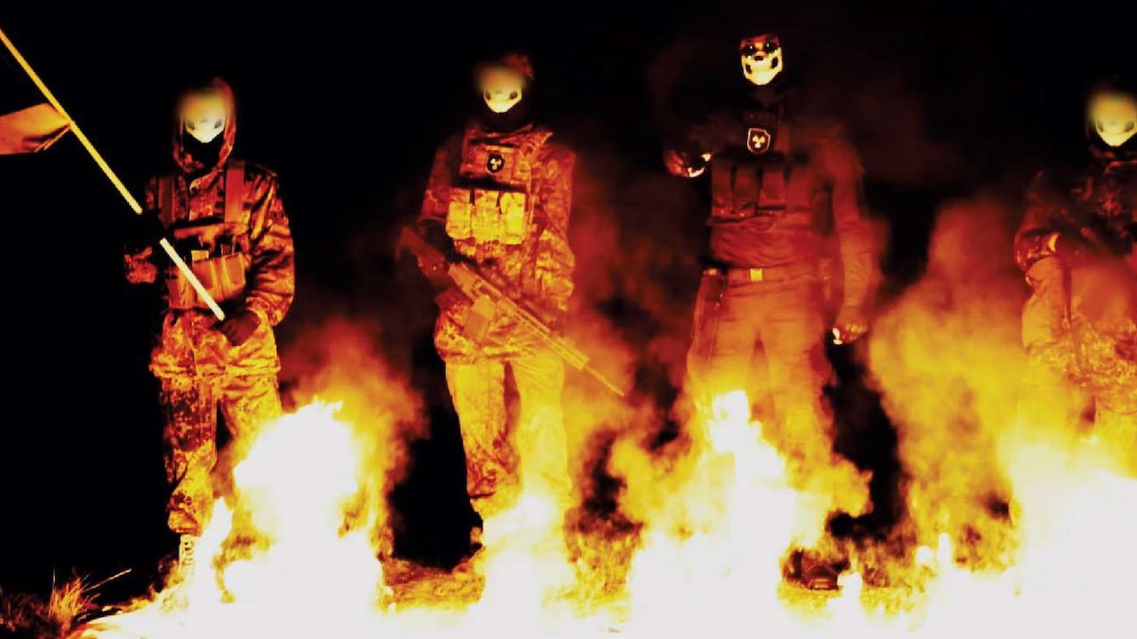 default-262 A group of men in masks and combat fatigues stand in front of a fire.
