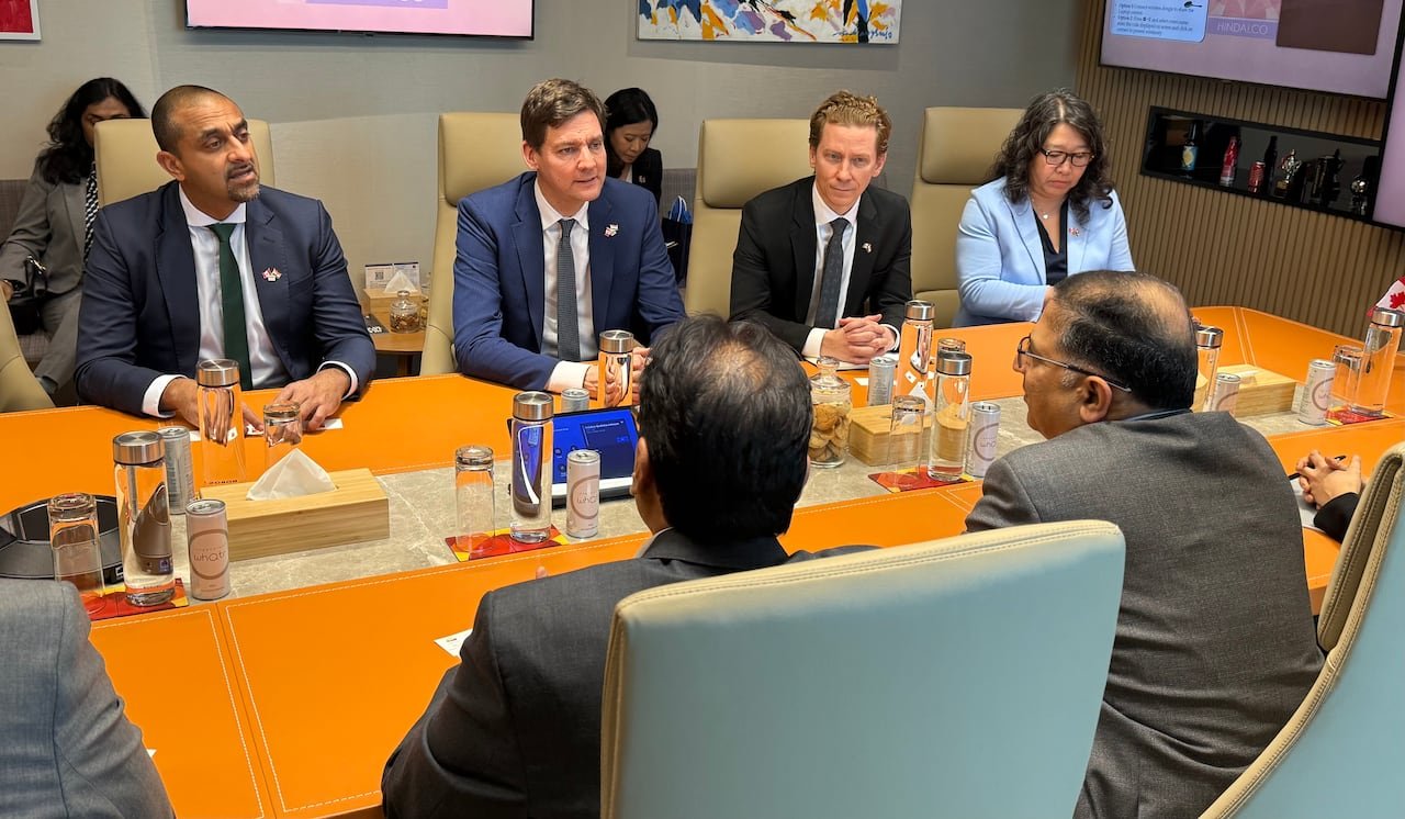 Three men and one woman sit on the far side of a table in a meeting. On the other side are two men in frame.