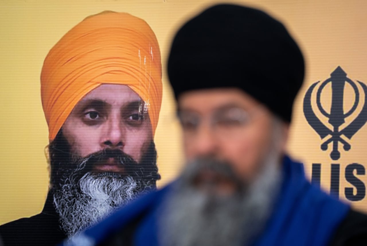 A photo of a man with a beard and orange turban is seen in the background. In front is a blurry man with a beard and black turban.